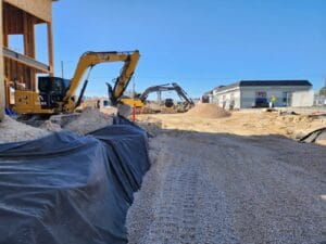 Construction site with two excavators working; a pile of gravel and mounds of dirt are visible. A worker in safety gear stands nearby, and a partially constructed building is on the left.
