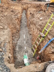 A trench showing a gravel bed with a green pipe and white markers. A yellow ladder leans against one side of the trench.