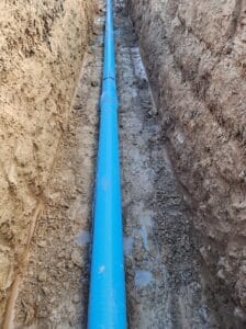 A blue pipe runs through a narrow trench dug in the earth, surrounded by brown soil.