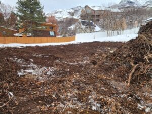 Cleared land with piles of dirt and debris, bordered by a wooden fence, with snow-covered mountains and houses in the background.