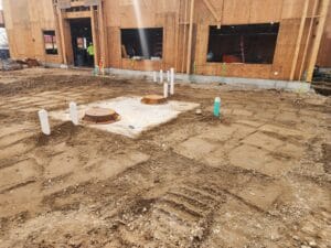 Construction site with exposed plumbing pipes and unfinished wooden structure. Dirt ground surface with cover openings in the center.