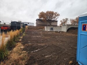 Construction site with cleared dirt area, a porta-potty on the right, parked vehicles, and buildings in the background under a cloudy sky.