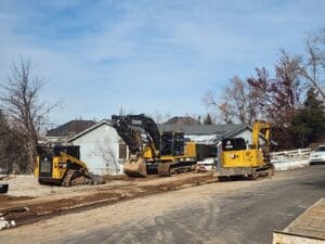 Construction site with three yellow excavators working on a dirt road. A residential house and bare trees are in the background under a cloudy sky.