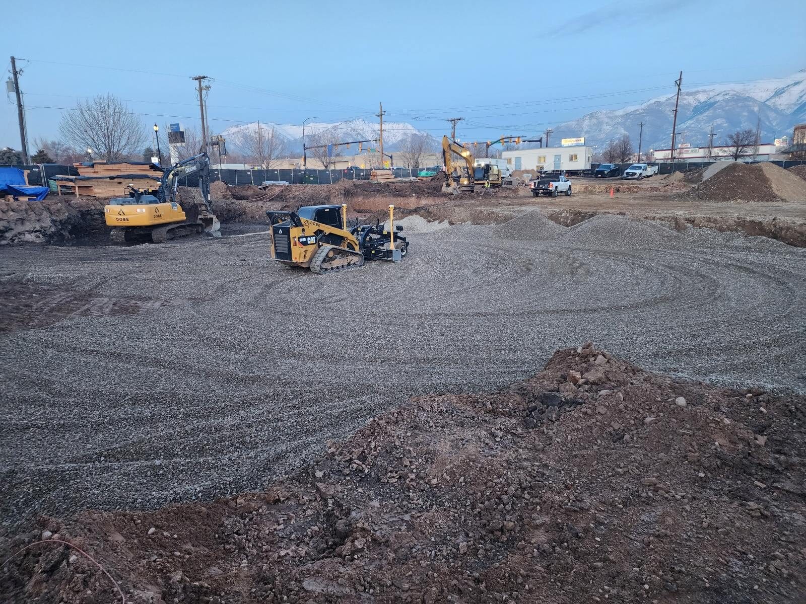 Construction site with several machines leveling gravel. Mountain and building in the background under a blue sky.