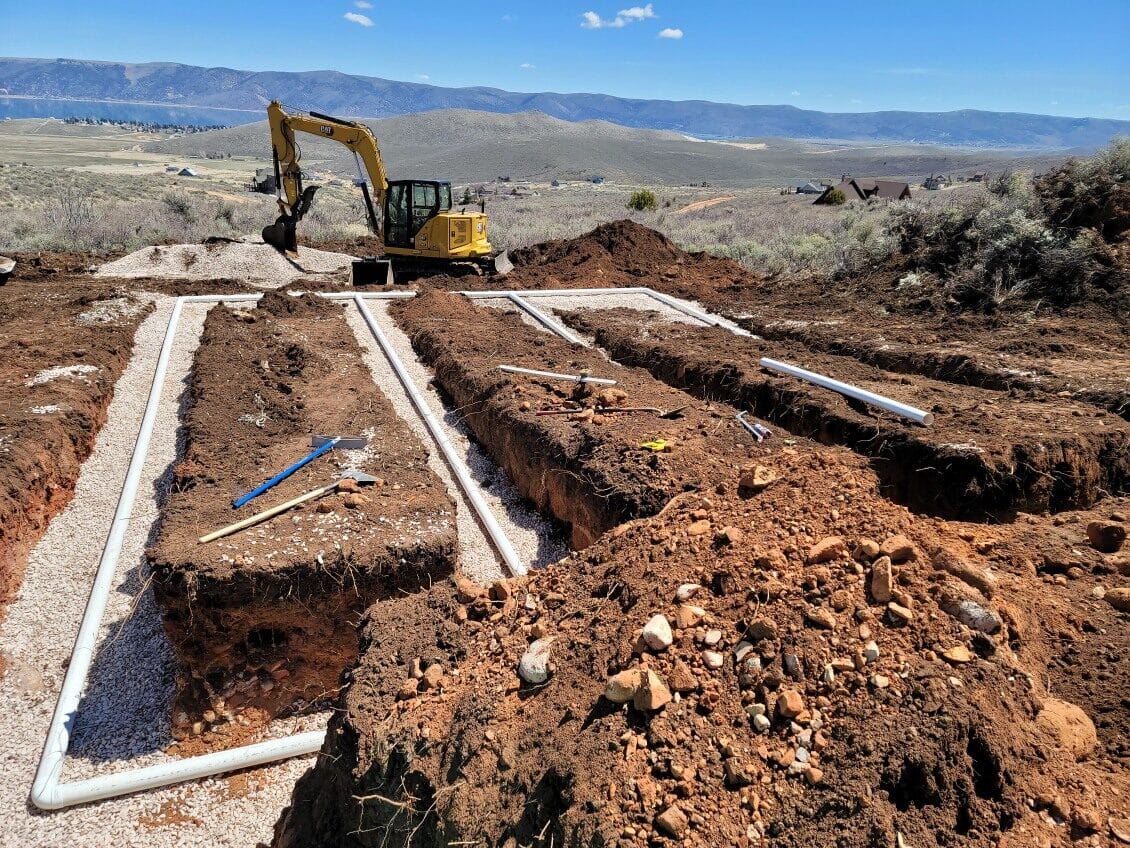 Excavator at a construction site with trenches and pipes laid in a rocky, rural landscape under a clear blue sky.