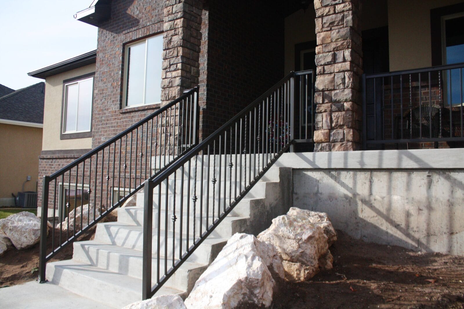 Concrete stairs with black metal railings lead to the entrance of a brick house. Large stones are placed beside the stairs.