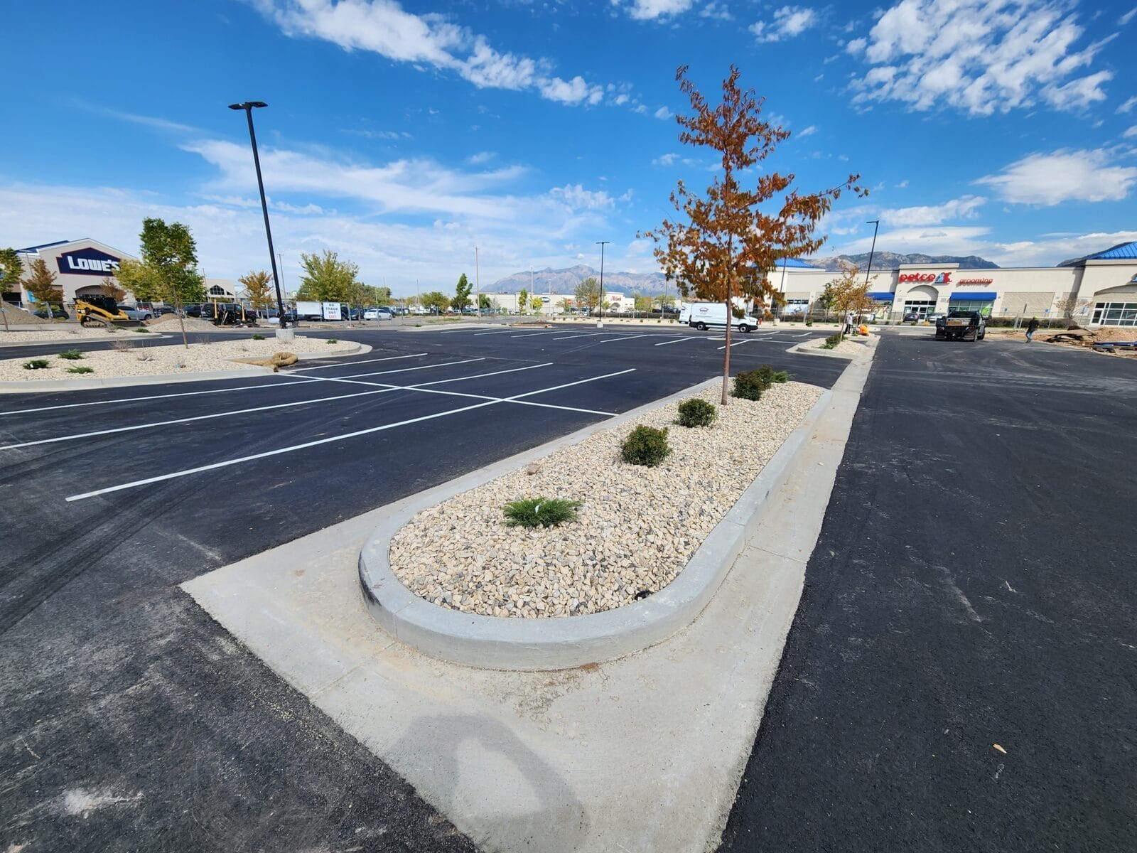 Parking lot with empty parking spaces and landscaped islands. Trees and small shrubs are planted. Stores, including a Lowe's, are visible in the background.