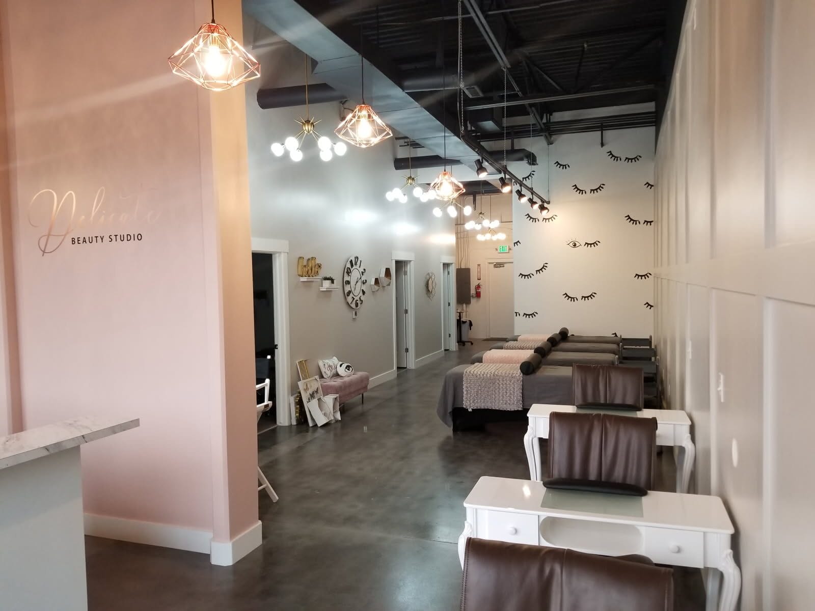 Modern beauty studio interior with pink and white walls, brown and white salon chairs, decorative wall art, and hanging ceiling lights. Tables are aligned along the right wall.