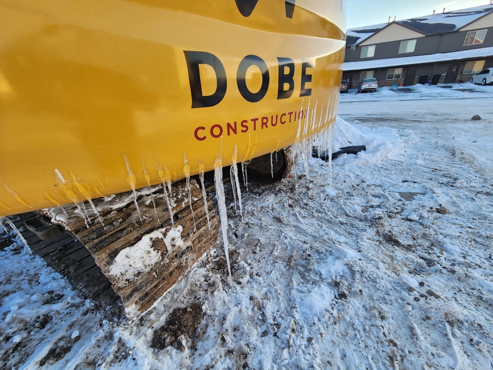 Excavator with icicles hanging from its tracks is parked on a snowy site near residential buildings.